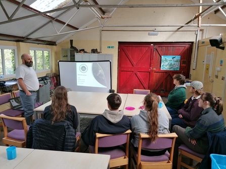 A group of people sit around a table as Gareth presents about the urban riverfly programme