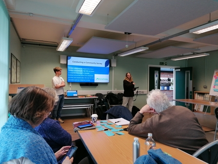 A man and a woman stand in front of a presentation screen, with a crowd of people watching