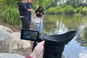 Three people stand near a river while a camera films them