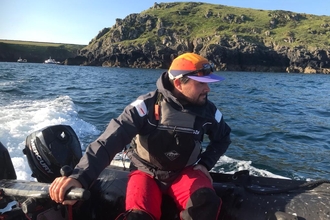 Leighton driving a boat around Skomer. The rocky island is in the background and the sea is a deep blue. 
