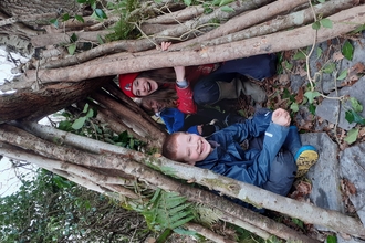 3 children sitting in a woodland  shelter made from branches