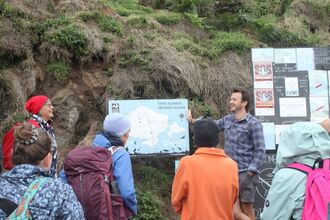 A guide points at the map with a crowd watching. There are signs behind them and a grassy bank.