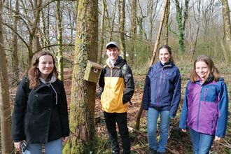 A group of people stood around a bird box on a tree, in a woodland.