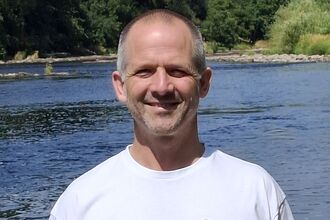 A man with short hair stood wearing a white t-shirt, stood in front of a lake surrounded by trees.