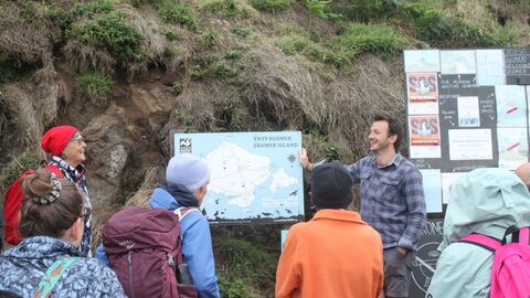 A guide points at the map with a crowd watching. There are signs behind them and a grassy bank.