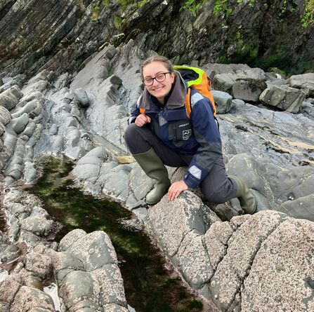 A woman crouched over a rockpool. She is smiling at the camera.