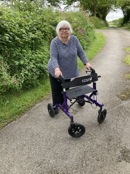 A woman stood with a walking aid on a path with rich green verges on each side. 