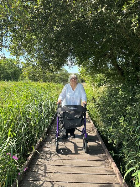 A woman stood with a walking aid on a boardwalk through a lush green nature reserve