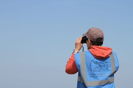 A person stood looking thorough binoculars, wearing a blue high-vis jacket.
