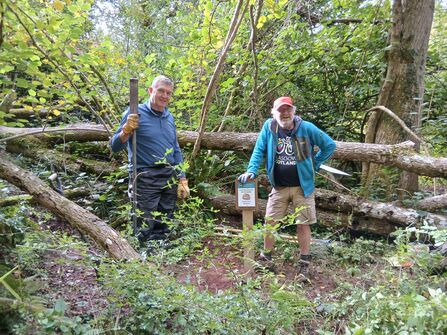 Two people stood next to a sign on a wooden post. They are surrounded trees, logs and vegetation.