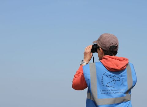 A person stood looking thorough binoculars, wearing a blue high-vis jacket.